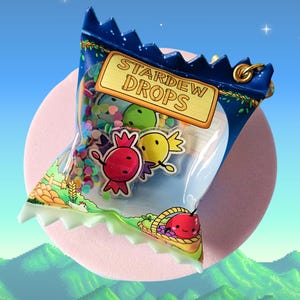 May include: A clear plastic bag of "Stardew Drops" sweets, with a blue and gold trim. The bag features colourful confetti and cartoon sweets in red, green, and yellow. The background is a pink circle with a pixelated landscape.
