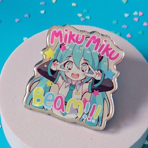 May include: A blue and pink anime character pin with the text "Miku Miku Beam!!" on a white background.