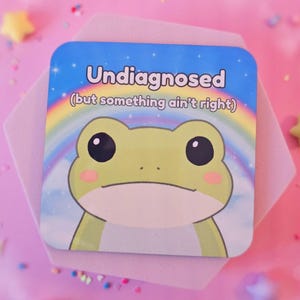 May include: A square coaster featuring a cartoon frog. The coaster has the text "Undiagnosed (but something ain't right)" above the frog, set against a blue sky and rainbow. The frog is light green with pink cheeks.