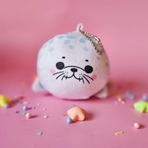 May include: A white plush seal toy with light blue spots and a silver chain. The seal has a smiling face with black eyes, whiskers, and pink cheeks. The background is pink, with colourful sprinkles.