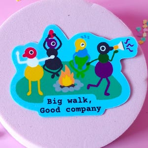 May include: A die-cut sticker featuring four colorful cartoon creatures gathered around a campfire on a green patch. One creature holds a megaphone, another sleeps, and two others sit. The text "Big walk, Good company" is printed in black.