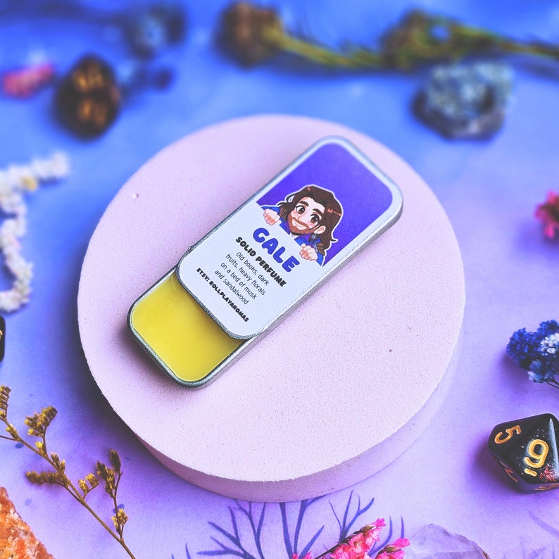 K&ouml;nnte beinhalten: Eine kleine, runde, silberne Dose mit einem gelben festen Parf&uuml;m darin. Die Dose zeigt eine Comic-Illustration einer Person mit braunen Haaren und einem blauen Hemd. Der Text auf der Dose lautet "GALE SOLID PERFUME Old books, dark fruits, heavy florals on a bed of musk and sandalwood ETEY: ROLLPLAYAROMAS".