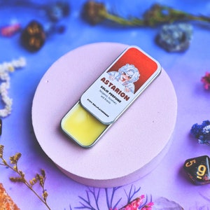 K&ouml;nnte beinhalten: Eine runde, rosafarbene Oberfl&auml;che mit einer kleinen, silbernen Dose, die ein festes, gelbes Parfum enth&auml;lt. Die Dose hat ein buntes Etikett mit dem Text "Astarion Solid Perfume Bay Leaf, Rosemary and Brandy" und einer Cartoon-Illustration einer Person.