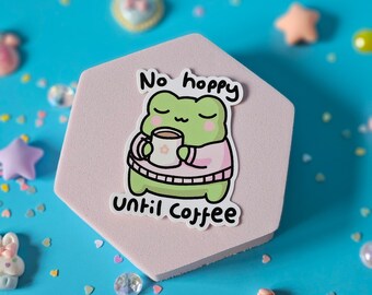 May include: A pink hexagonal coaster featuring a green frog wearing a pink sweater and holding a cup of coffee. The text "No hoppy until coffee" is printed on the coaster.