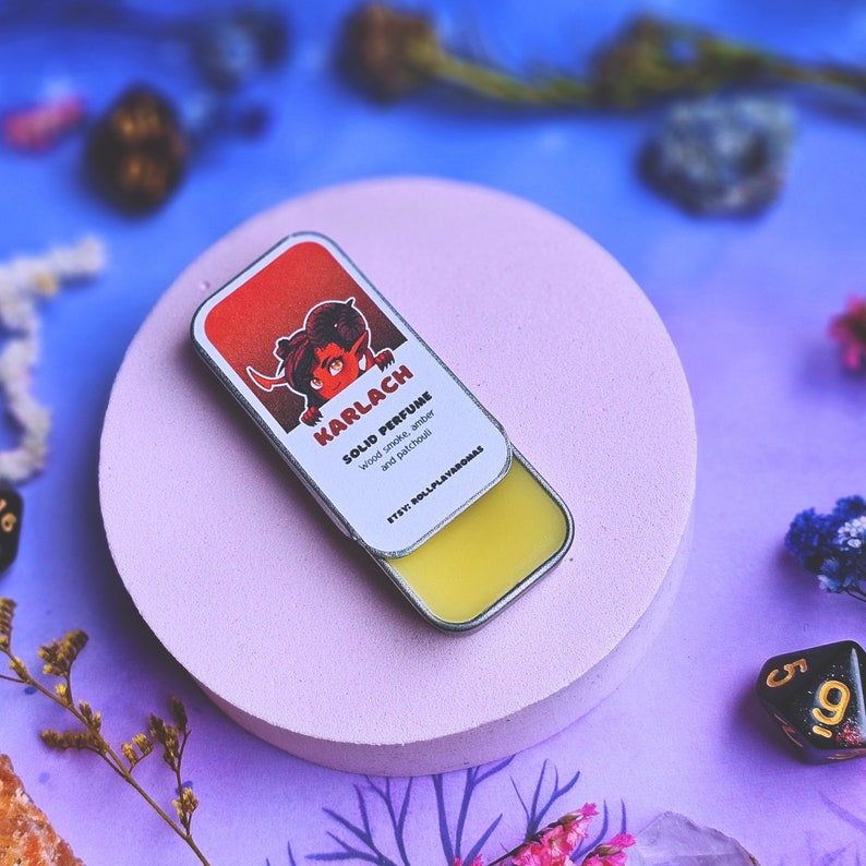 K&ouml;nnte beinhalten: Eine kleine, runde, silberne Dose mit einem roten und schwarzen Etikett, auf dem "KARLACH SOLID PERFUME wood smoke, amber and patchouli ETSY: ROLLPLAYAROMAS" steht. Die Dose ist ge&ouml;ffnet und zeigt ein gelbes festes Parf&uuml;m im Inneren.