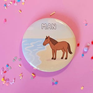Puede incluir: Un botón redondo con la palabra "MAN" en letras negras en la parte superior. Debajo, un caballo marrón de pie en una playa de arena con agua azul y un cielo azul claro de fondo.