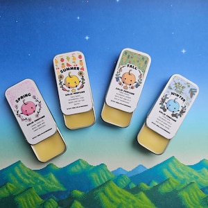 K&ouml;nnte beinhalten: Vier kleine runde Dosen mit bunten Deckeln und Etiketten. Jede Dose zeigt eine andere Jahreszeit: Fr&uuml;hling, Sommer, Herbst und Winter. Jede Dose hat einen Comic-Apfel mit einer anderen Farbe und einem anderen Ausdruck. Der Text auf den Dosen lautet "Festes Parf&uuml;m" und listet die D&uuml;fte f&uuml;r jede Jahreszeit auf.