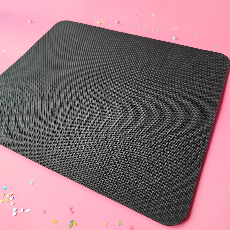 May include: A black mouse mat with a textured surface. The mouse mat is on a pink background.