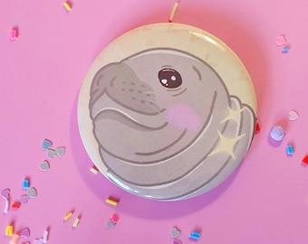 May include: A round button featuring a cartoon illustration of a gray seal with a light yellow background. The seal has a cute expression with pink cheeks and a starburst design. The button is on a pink surface with colorful confetti.