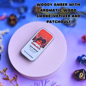 K&ouml;nnte beinhalten: Eine kleine, silberne Dose mit einem roten und wei&szlig;en Etikett, auf dem "KARLACH SOLID PERFUME wood smoke, amber and patchouli" steht und eine Comic-Illustration einer rot-h&auml;utigen Person mit H&ouml;rnern und einem Schwanz zu sehen ist. Die Dose steht auf einer runden, rosafarbenen Oberfl&auml;che.