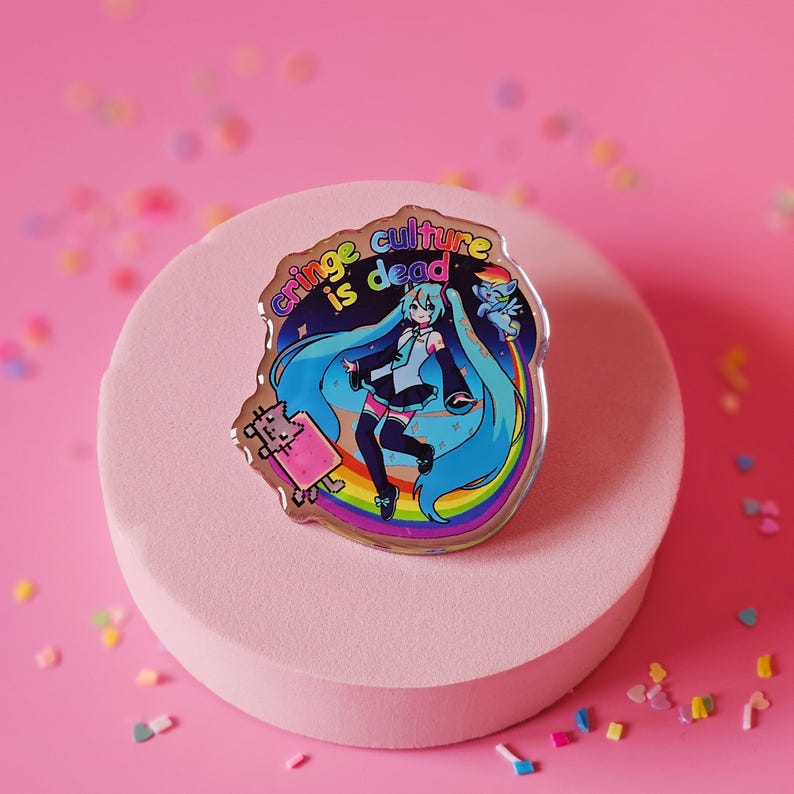 Op de afbeelding: Een regenboog holografische pin met de tekst "cringe culture is dead" en een cartoon afbeelding van een anime-personage met blauw haar en een regenboog erachter.