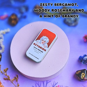 K&ouml;nnte beinhalten: Eine silberne Dose mit einer Cartoon-Illustration einer Person mit wei&szlig;en Haaren und roten Augen. Die Dose ist mit "Astarion Solid Perfume Bergamot, Rosemary and Brandy" und "Etsy: RollPlayAromas" beschriftet.
