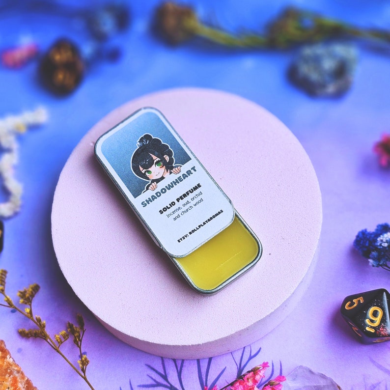 K&ouml;nnte beinhalten: Eine kleine, silberne Dose mit einem gelben festen Parf&uuml;m darin. Die Dose zeigt eine Comic-Illustration eines M&auml;dchens mit gr&uuml;nen Augen und schwarzen Haaren. Der Text auf der Dose lautet "SHADOWHEART SOLID PERFUME Incense, oud, orchid and church wood ETSY: ROLLPLAYAROMAS".