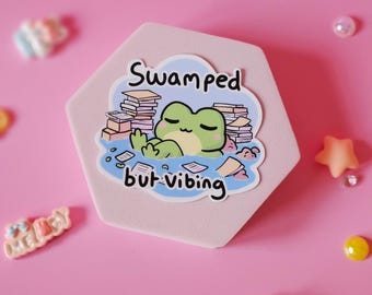 May include: A hexagonal sticker featuring a cartoon frog surrounded by papers and books. The text on the sticker reads "Swamped but-vibing".