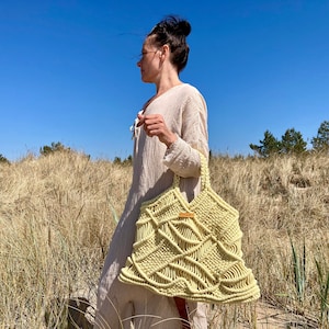 Peut inclure: Une femme marche dans un champ d'herbe haute portant un grand sac fourre-tout en macramé jaune clair avec une poignée en cuir marron.