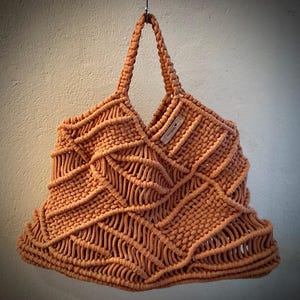 May include: A large, brown, crocheted tote bag with a woven pattern. The bag has a single strap and a small, brown leather tag with the word "Handmade" on it.