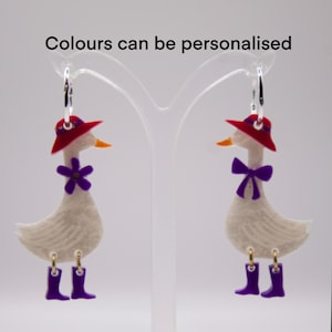 May include: A pair of dangle earrings featuring white goose charms wearing red hats and purple boots. One goose has a purple flower at the neck, while the other has a purple bow. The text "Colours can be personalised" is visible above.