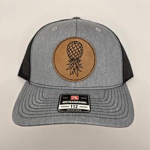 May include: A gray and black trucker hat with a brown leather patch featuring a pineapple design. The hat is adjustable and has the text "Richardson. 112" on the front.