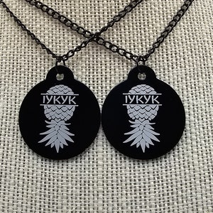 May include: Two black necklaces with round black pendants featuring a white pineapple design and the text "IYKYK".