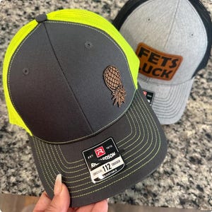 May include: Two trucker hats, one gray with a brown leather patch that says "FETS LUCK" and the other gray with a neon green mesh back and a brown pineapple patch. The hat with the pineapple patch has a black and white label that says "EST. 1970 RICHARDSON ORIGINAL 112 TRUCKER ADJUSTABLE".