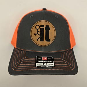 May include: A black and orange trucker hat with a brown leather patch that says "It" with a stick figure flipping the bird. The hat is adjustable and has the Richardson 112 logo on the back.