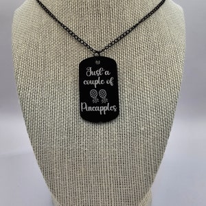 May include: A black necklace with a black metal dog tag pendant. The pendant has the text "Just a couple of Pineapples" engraved on it.