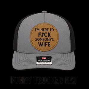 May include: A grey and black trucker hat featuring a circular tan patch on the front. The patch displays the black text I'M HERE TO F*CK SOMEONE'S WIFE. A small rectangular black label with the white text RICNASOSOM is centered on the brim.
