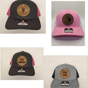 May include: Four trucker hats with different patches. The first hat is black with a pink mesh back and a brown leather patch that says "Plays Well With Others" and has a pineapple design. The second hat is pink with a white mesh back and a brown leather patch that says "Pussycat Parties". The third hat is black with a pink mesh back and a brown leather patch that says "I'm Here To Fick Someone's Husband". The fourth hat is gray with a black mesh back and a brown leather patch that says "I'm Here To Fick Someone's Wife".