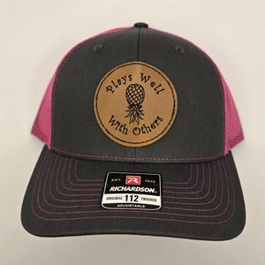 May include: A gray and pink trucker hat with a brown leather patch featuring a pineapple and the words "Plays Well With Others." The hat has a curved brim and a mesh back. The Richardson brand label is visible.