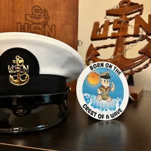 Puede incluir: Un sombrero blanco de la Marina de los Estados Unidos con un emblema de ancla dorada y una banda negra. Junto a él hay una pegatina redonda con un marinero de dibujos animados en una ola y el texto "Born on the Crest of a Wave".