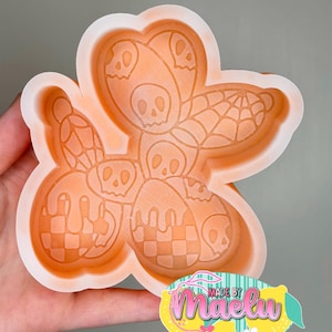 May include: An orange silicone mold in the shape of a flower with a skull and spiderweb design. The mold features a checkerboard pattern and skull details. The mold is held in a hand, with a "Made by Maelu" sticker in the corner.