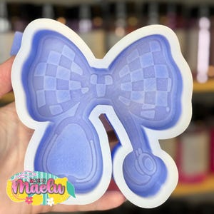 May include: A light purple silicone mold in the shape of a bow with a checkered pattern. The mold has a white border and a design of a bow and two teardrop shapes. The text "Made by Maelu" is visible in the lower left corner.