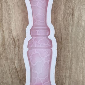 May include: A pink silicone mold in the shape of a vase. The interior of the vase has a pattern of irregular shapes. The mold is outlined in white and sits on a wooden surface. The mold is designed for crafting.