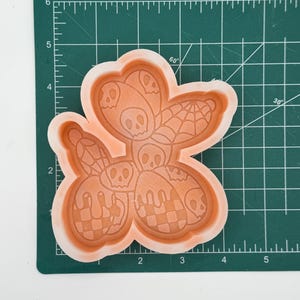 May include: An orange silicone mold with a Halloween theme. The mold features a flower shape with skull, spiderweb, and checkered designs. The mold is on a green cutting mat with measurement markings.