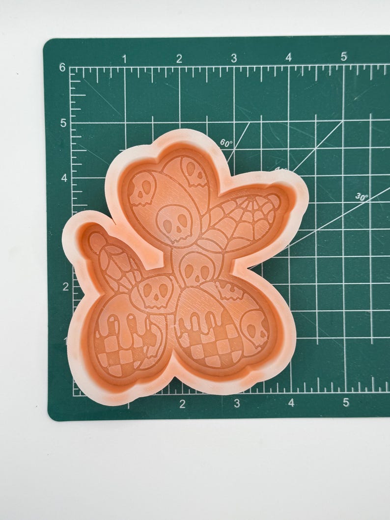 May include: An orange silicone mold with a Halloween theme. The mold features a flower shape with skull, spiderweb, and checkered designs. The mold is on a green cutting mat with measurement markings.