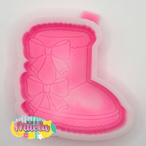 May include: A pink, plastic boot-shaped mold with two large bows and a fluffy top. The mold is outlined in a lighter pink and has the text "Made by Maelyn" in a colorful logo.