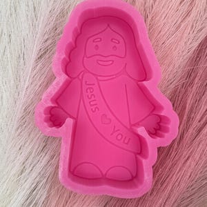 May include: A bright pink silicone mold in the shape of Jesus with a banner that reads "Jesus You". The mold is designed with a smiling face and outstretched arms, perfect for crafting projects.
