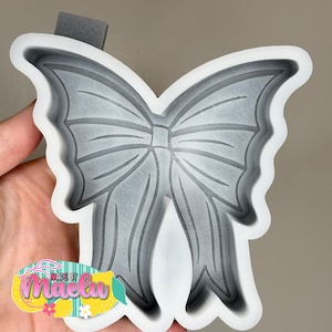 May include: A gray silicone mold in the shape of a bow. The mold has detailed lines to create the bow's shape. The mold is held by a hand, and a sticker with the text "Made by Maelu" is visible.
