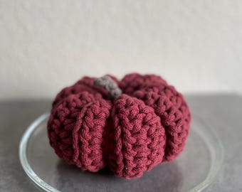 May include: A crocheted burgundy pumpkin with a gray stem. The pumpkin is sitting on a clear glass plate.