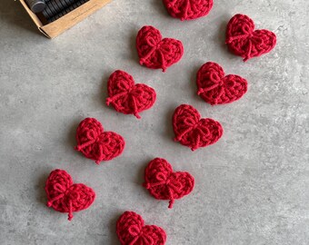May include: A collection of red crocheted heart-shaped ornaments, each with a bow detail. A small cardboard box filled with cylindrical magnets is visible in the background. The hearts are scattered on a gray surface.