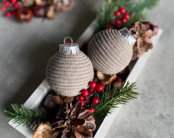 May include: Two brown crocheted Christmas ornaments with silver tops in a white tray with pine sprigs, pine cones, and red berries.