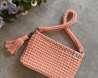 May include: A peach-colored crocheted bag with a long strap and a tassel. The bag is made of a thick, textured yarn and has a simple, minimalist design.