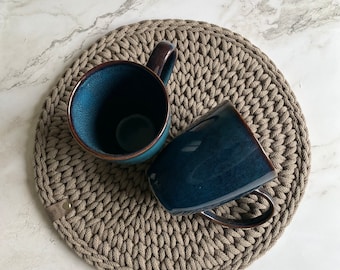 May include: Two dark blue ceramic mugs with brown rims sit on a beige crocheted coaster. The coaster has a brown leather strap.