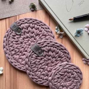 Könnte beinhalten: Ein Set aus drei handgefertigten, runden, mauvefarbenen Häkeluntersetzern. Jeder Untersetzer hat ein strukturiertes Muster und ein kleines Lederetikett mit dem Wort "handmade". Ein Notizbuch mit dem Wort "Create 2024" und ein Stift befinden sich im Hintergrund.