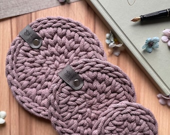 May include: Three circular, mauve-colored crocheted coasters of varying sizes, each featuring a thick, braided yarn texture. Two of the coasters are adorned with a small, dark brown leather-like tab secured by a silver metal rivet.
