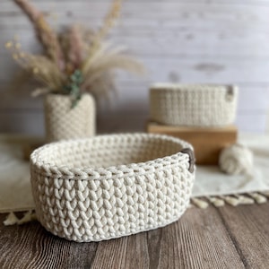 May include: Cream-colored, oval-shaped crochet basket with a leather accent. The basket is made of thick yarn and sits on a wooden surface. A smaller basket and a vase with dried flowers are in the background.