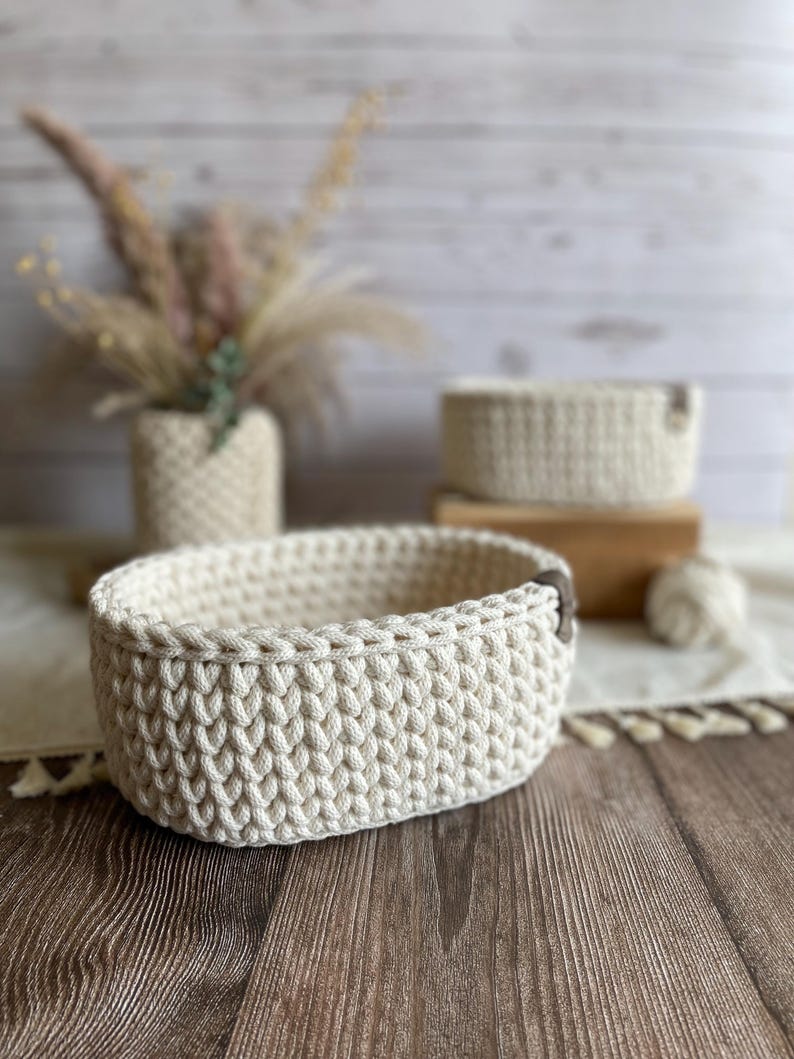 May include: Cream-colored, oval-shaped crochet basket with a leather accent. The basket is made of thick yarn and sits on a wooden surface. A smaller basket and a vase with dried flowers are in the background.