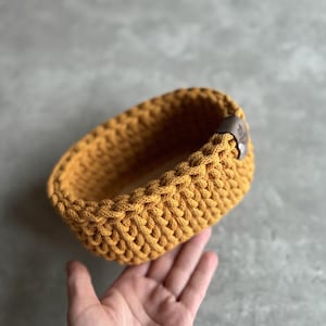 May include: Mustard yellow crochet basket with a braided design. The oval-shaped basket has a brown leather tag attached to the side. The basket is handmade and suitable for storage or decoration.