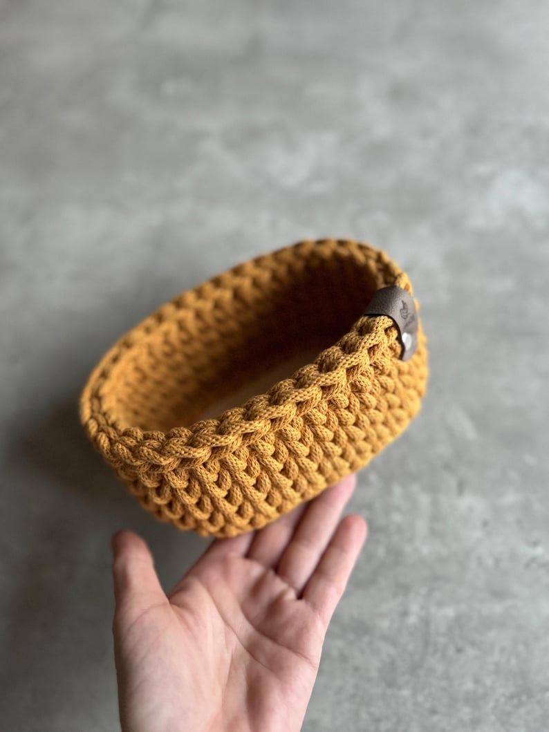 May include: Mustard yellow crochet basket with a braided design. The oval-shaped basket has a brown leather tag attached to the side. The basket is handmade and suitable for storage or decoration.