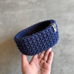 May include: A navy blue, hand-crocheted basket with a rounded rectangular shape. The basket is made of thick, textured yarn and features a small, dark leather tag. The basket is held in a hand against a gray background.